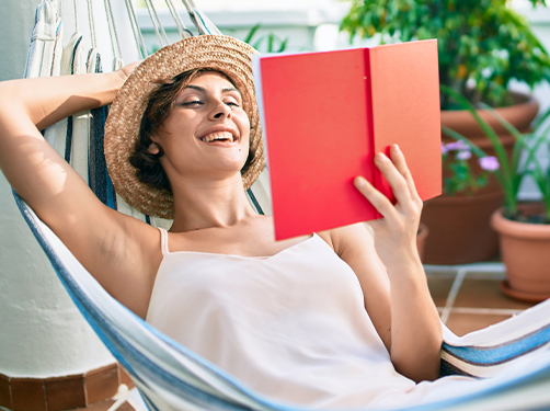 Frau liegt entspannt in der Hängematte und liest ein Buch. Grüne Pflanzen im Hintergrund vervollständigen die sommerliche Atmosphäre.